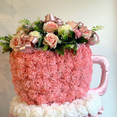 Tea Rose Tribute - A unique and personal funeral tribute in the shape of a teacup, adorned with soft pink blooms and decorative ribbons. This design brings a comforting and nostalgic touch to a memorial service.